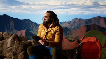 Camper fighting the strong wind while he uses a tablet at a high altitude campsite, searching for better signal and risking his safety. Dangerous mountain range cold weather. Camera B. video