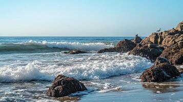 A serene beach scene with waves crashing on rocks under a clear blue sky photo