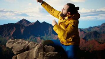 Determined adventurer enjoys the panoramic mountain landscape, taking selfies with the amazing scenic background on the hill top where nature and towering peaks create a breathtaking view. Camera A. video