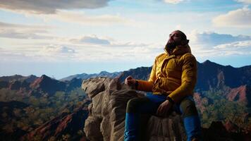 Male mountain hiker celebrates a victorious climb and resting at the top, arms lifted in excitement while he sits on the rocks. Panoramic view and crisp air in a scenic background. Camera A. video