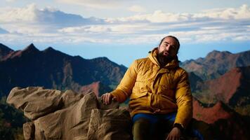 Satisfied pleased hiker reaching the top of the mountain and being tired, resting and relaxing on the rocky mountain peak. Young man enjoying the breathtaking scenic view. Camera B. video