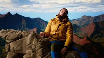 Male mountain hiker celebrates a victorious climb and resting at the top, arms lifted in excitement while he sits on the rocks. Panoramic view and crisp air in a scenic background. Camera B. video