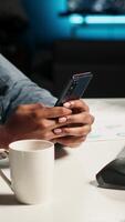 Vertical African american man listening to music and browsing webpages on his mobile phone, scrolling through social media apps and taking break from solving tasks at the desk. Answering texts. Camera B. video