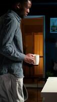 vertical yo empleado negro hombre sentado a el escritorio organizando su cronograma, revisando en línea datos notas chico con un café taza hojeada el Internet para investigación, remoto trabajo en un moderno desván. cámara una. video