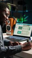 Vertical Black freelancer researching online data and writing notes for articles, sitting at his personal home office. Young man working from home, balancing tasks using a wireless setup. Camera B. video