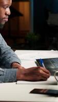 Vertical Self employed Black man multitasking at home taking handwritten notes and browsing the web for project research. Managing schedule and tasks with a planner to boost productivity. Camera B. video
