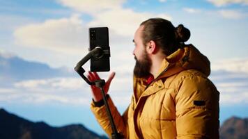Traveler filming his hiking experience in the wilderness documenting his adventure on social media using his smartphone. Recording the beautiful scenic mountain view behind him for content. Camera B. video