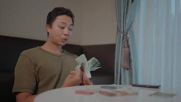 Counting cash at home. A young man sits at a table, meticulously counting a stack of banknotes. The scene is intimate and suggests financial planning or a moment of reflection on earnings. video