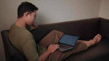 Cozy work from home a young man comfortably works on his laptop while relaxing on a sofa, showcasing a relaxed yet productive work-from-home atmosphere. video