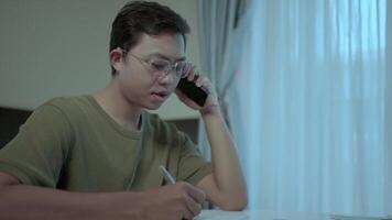 Focused young man on a call. A young man, wearing glasses, sits at a table, attentively taking notes while on a phone call. video