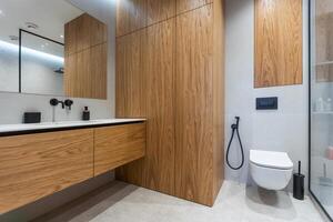 Bathroom in a modern style with gray and white tiles. There is a large mirror with luminous lamps, tabletop with wooden drawers and sink, bath with shower and glass partition, towel rack and a hanger. photo