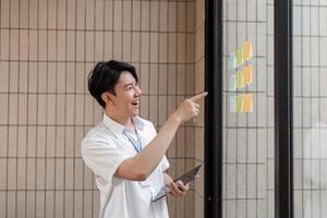 Young professional excitedly pointing at sticky notes on glass wall, discussing ideas and enhancing workflow in modern office. photo