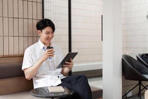 Young Asian male office worker smiling while using tablet in modern workspace, enjoying coffee and digital content. photo
