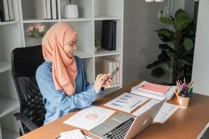 Muslim woman smiling while using smartphone in a modern office with financial documents. photo