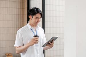 Young Asian male office worker standing and smiling while using tablet in modern workspace, enjoying coffee and digital interaction. photo