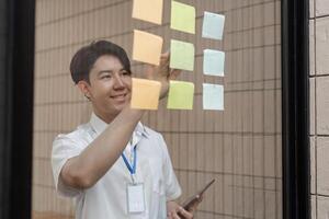 Young professional smiling while arranging sticky notes on glass wall, organizing tasks and improving workflow in modern office. photo