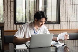 Young professional analyzing documents while working on laptop in modern office, focusing on data and project details. photo
