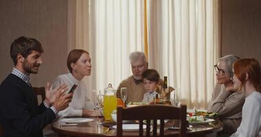 uma família sentado por aí uma mesa com uma homem e mulher usando uma célula telefone video