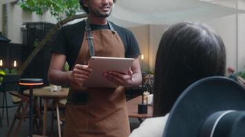 Smiling cafe worker waiter man barista chef in apron talk customer order food lunch dinner in restaurant Indian Arabian latino guy serving visitor in coffee shop client service using digital tablet video