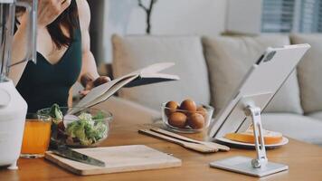 A woman is reading a book while sitting at a table with a laptop and food video