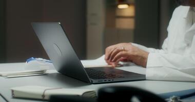 The doctor is typing on a laptop and writing in a notebook sitting at a table in a medical office, working in a clinic. Close-up of his hands, an unrecognizable person video
