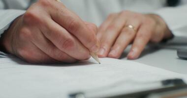 A doctor in a white coat is writing in a notebook sitting at a table, close-up of his hands, an unrecognizable person video