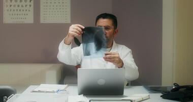 A doctor in a white coat examines an X-ray of a patient sitting in front of an open laptop at a table in a medical office, working in a clinic video