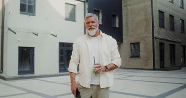 uma grisalho homem com uma bigode, e barba parece às a Câmera com uma sério Veja e bebidas limonada a partir de uma descartável copo com uma Palha em pé em uma cidade rua video