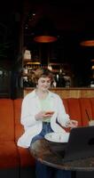 A woman uses a mobile phone and drinks coffee sitting at a table in a cafe in front of an open laptop video