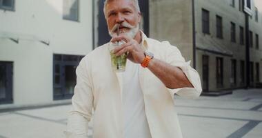 A gray-haired man with a mustache, and beard looks at the camera with a smile and drinks lemonade from a disposable cup with a straw standing on a city street video