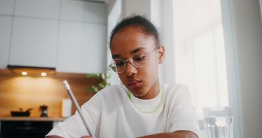 A girl writes in a notebook, doing homework sitting at the kitchen table at home, a close-up of her face video