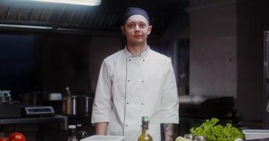 A man in a chef's uniform with a headband smiles while looking at the camera standing in a well-equipped restaurant kitchen video