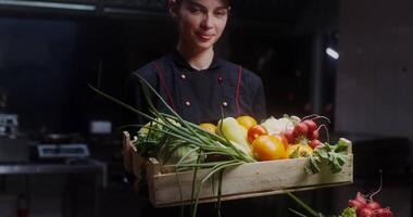 A young woman in a chef's uniform with a box of vegetables in her hands smiles while looking at the camera video