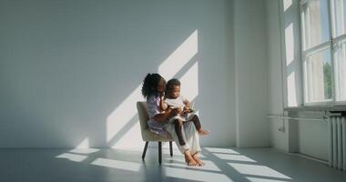 Mom with a child of African-American appearance, dressed in white clothes, playing pyramid while sitting in a chair in a white room near the window video