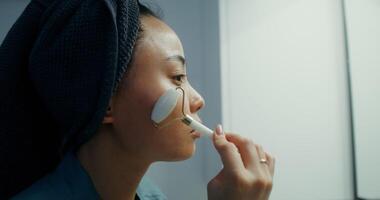 Young Asian woman with a towel around her head, doing a face massage using a roller massager, close-up of her face, side view video