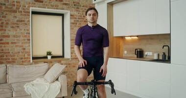 A young man of European appearance of an athletic build looks at the camera while sitting on an exercise bike in a stylish home interior video
