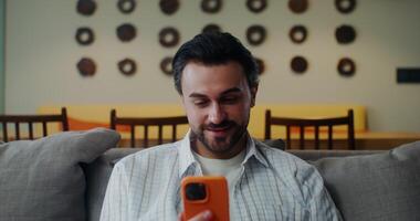 A man in casual clothes smiles and waves hello starting a call on a mobile phone while sitting on a sofa in a stylish home interior. Front view video