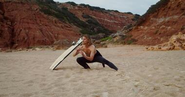 A young man in a diving suit with the top off does exercises standing on a sandy beach, next to a surfboard stuck in the sand video