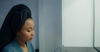 A young woman in a bathrobe, with a towel around her head, applies a moisturizer with a pipette to her face and rubs it with her hands, looking in the mirror in the bathroom, close-up video