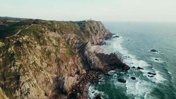 The drone flies along the seashore with rocks, the tops of which are covered with grass. The lighthouse stands on the top of a mountain in the distance video