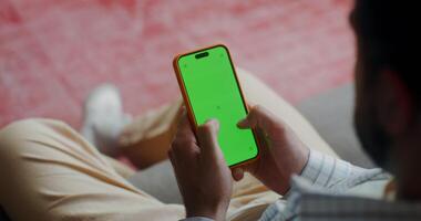 A man in casual clothes typing on a mobile phone with green screen sitting on a sofa indoors. Top view, no face, close-up of a man's hands video