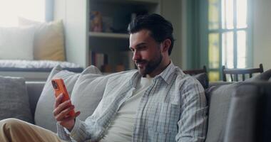 A man in casual clothes smiles, and waves hello starting a call on a mobile phone while sitting on a sofa in a stylish home interior video