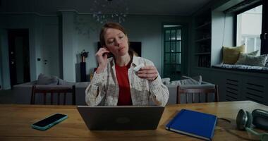 A red-haired woman puts on headphones while talking on a chat on a laptop, sitting at a table in a stylish home interior with a serious look, working remotely from home video