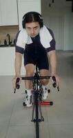 A young man in headphones with a towel around his neck looks at the camera while using an exercise bike, exercising in a modern home interior, vertical video