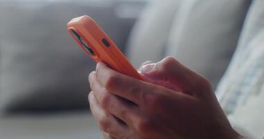 Close-up of a mobile phone with three cameras, in an orange case in men's hands indoors. Man typing on smartphone, no face video