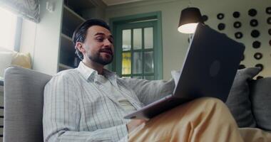 A stylish bearded man smiles and waves hello while talking on a call on a laptop standing on his lap, sitting on a sofa indoors video