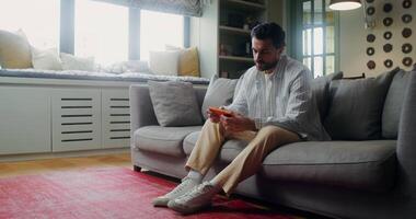 A young bearded man is playing a game on a mobile phone while playing sitting on the couch at home video