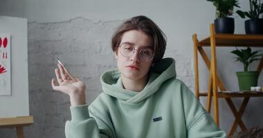 Young woman artist with a drawing brush in her hands looks at the camera with a serious look, sitting at the desktop in the workshop video