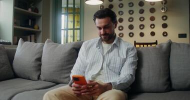 A man in casual clothes laughs while looking at the mobile phone screen while sitting on the couch at home video