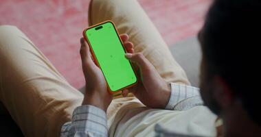 uma homem dentro casual roupas usa uma Móvel telefone com uma verde tela enquanto sentado em a sofá às lar. topo visualizar, não face video